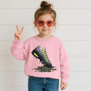May include: A pink sweatshirt featuring a graphic of a gray and black sneaker with neon green accents. The sneaker appears to be dripping a green liquid. The sweatshirt is worn by a young child wearing heart-shaped sunglasses.