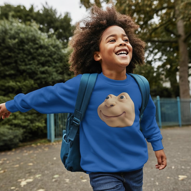 May include: A blue sweatshirt with a smiling frog face graphic. The sweatshirt is a solid blue colour and is being worn by a child with a backpack. The child is smiling and appears to be outside.