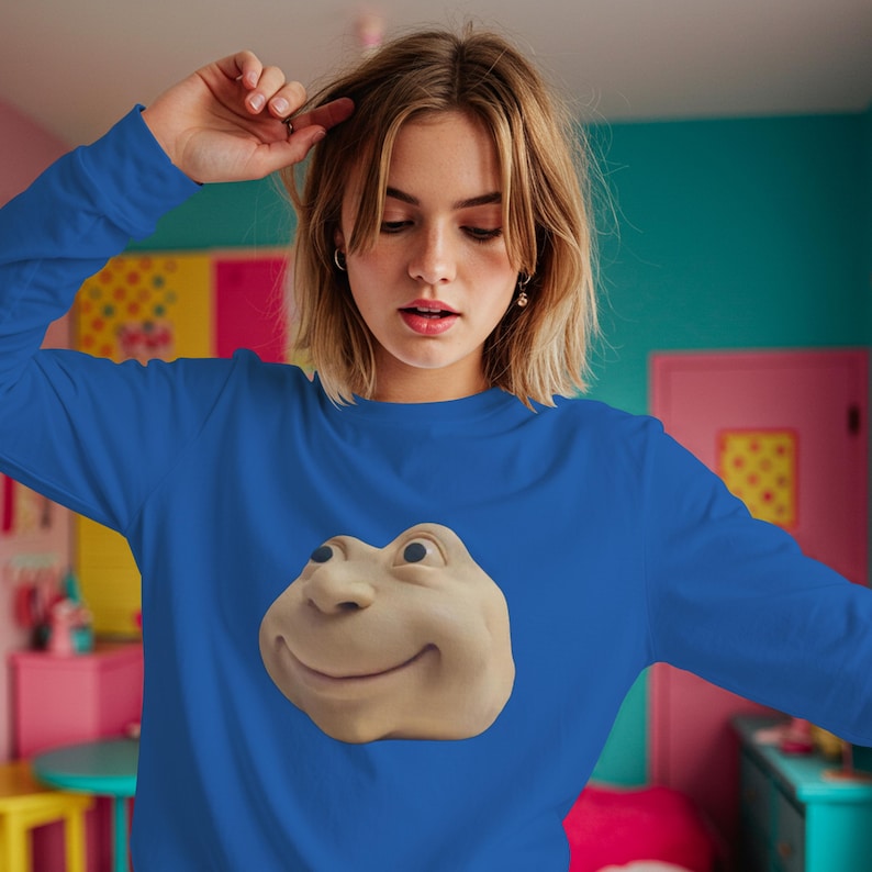 May include: Royal blue long-sleeve top with a smiling, beige cartoon face graphic on the front. The graphic has a nose, eyes, and a smiling mouth. The top is made of a soft material.