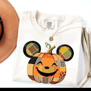 May include: Cream-colored Comfort Colors t-shirt featuring a smiling pumpkin design with Mickey Mouse ears. The pumpkin is orange with patchwork details in various colors and patterns. A gold necklace and a brown hat are also visible.