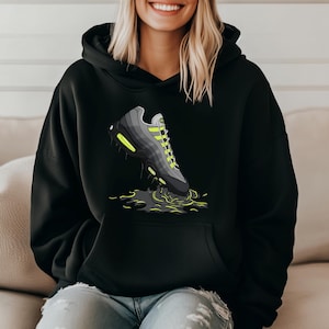 May include: Black hoodie featuring a graphic of a gray and black sneaker with neon green accents. The sneaker is depicted in a puddle of dark green liquid. The hoodie has a front pocket and a hood.