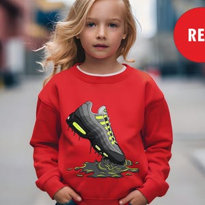 May include: A red sweatshirt featuring a graphic of a sneaker with black, gray, and neon yellow accents. The word "RED" is in a red circle in the upper right corner. The sweatshirt is worn by a young person.