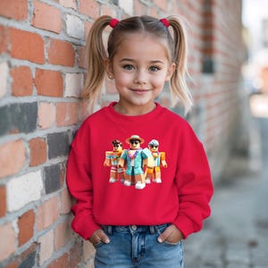 Puede incluir: Una ni&ntilde;a peque&ntilde;a con una sudadera roja brillante con un gr&aacute;fico de tres personajes de dibujos animados con ropa de playa. Los personajes llevan gafas de sol, sombreros y camisas hawaianas. La ni&ntilde;a tambi&eacute;n lleva vaqueros azules.