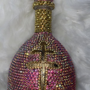 May include: A decorative bottle covered in iridescent rhinestones. The bottle is primarily pink and gold, with a gold cross design. The bottle's neck and stopper are also adorned with rhinestones, creating a luxurious and eye-catching design.