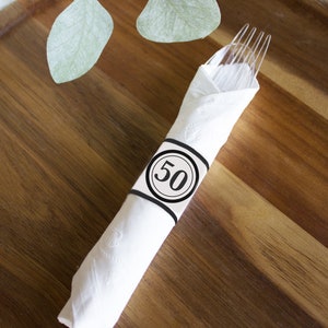 Könnte beinhalten: Eine weiße Serviette mit einem schwarz-weißen Serviettenring mit der Nummer 50. Die Serviette ist um eine transparente Plastikgabel gewickelt. Serviette und Gabel liegen auf einer Holzoberfläche.