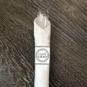Könnte beinhalten: Weiße Serviette mit einem grauen und weißen Kreis, auf dem "congrats grad" steht, um eine Gabel und einen Löffel herum.