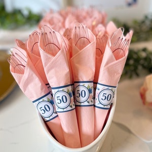 Könnte beinhalten: Eine weiße Keramikschale gefüllt mit rosa Servietten, jede um eine transparente Plastikgabel gewickelt. Die Servietten sind mit einem dekorativen Band mit der Zahl "50" in einem floralen Design verziert, für eine Feier.