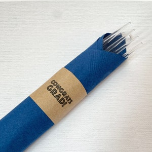 Könnte beinhalten: Ein Satz durchsichtiger Glas-Trinkhalme, verpackt in blaues Papier mit einem braunen Etikett, auf dem "Congrats Grad!" steht.