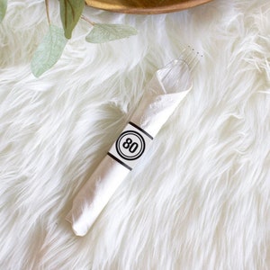 Könnte beinhalten: Eine weiße Serviette mit einem schwarzen und weißen Kreis mit der Nummer 80 darauf. Die Serviette ist um eine Gabel gewickelt.