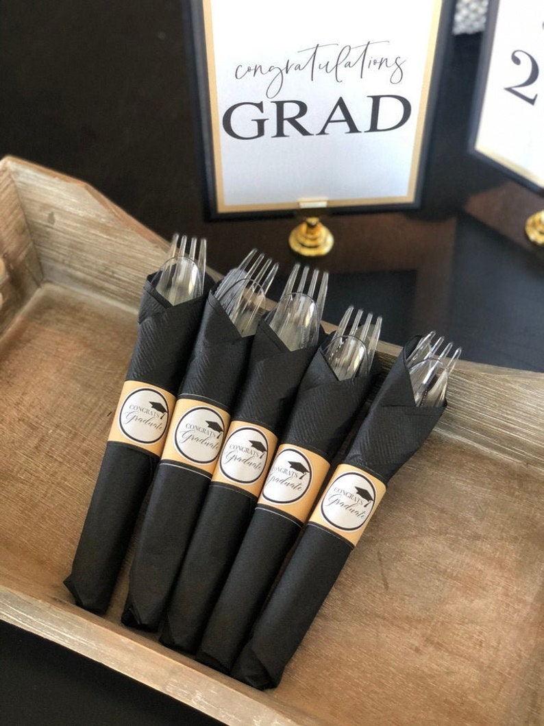 Puede incluir: Servilletas negras con pegatinas doradas de gorro de graduaci&oacute;n que dicen "Congrats Graduate" envueltas alrededor de tenedores de pl&aacute;stico transparentes.