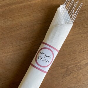 Könnte beinhalten: Weiße Serviette mit einem rosa und weißen Kreis, auf dem "congrats GRAD" steht. Die Serviette ist aufgerollt und enthält ein Set durchsichtiger Plastikgabeln.