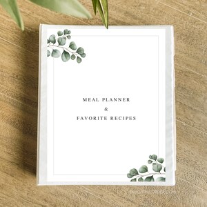 Puede incluir: Carpeta blanca con un dise&ntilde;o de hojas verdes en la portada. El texto "MEAL PLANNER & FAVORITE RECIPES" est&aacute; impreso en negro en la parte delantera.