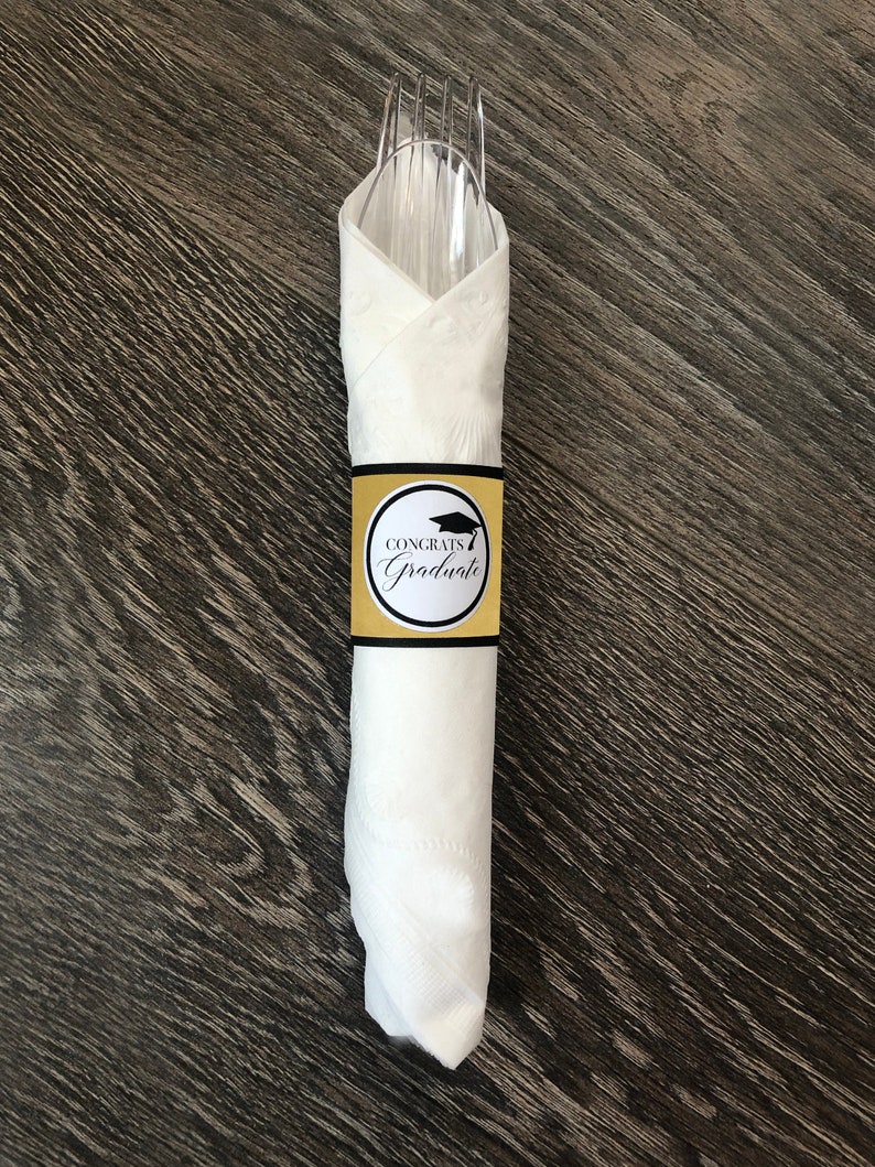 Puede incluir: Servilleta blanca con un anillo de servilleta de gorro de graduaci&oacute;n dorado y negro que dice "Congrats Graduate".