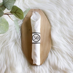 Könnte beinhalten: Weiße Serviette mit einem schwarz-weißen Ring mit der Nummer 50 auf einem Holzbrett. Das Brett liegt auf einer weißen, flauschigen Oberfläche.