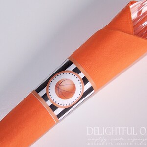 May include: Orange napkin with a black and white striped band with a basketball graphic. The band has a white circle with black polka dots around the basketball.