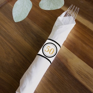 Könnte beinhalten: Eine weiße Serviette mit einem schwarzen und goldenen Serviettenring. Der Serviettenring hat einen Kreis mit der Zahl 50 in Gold.