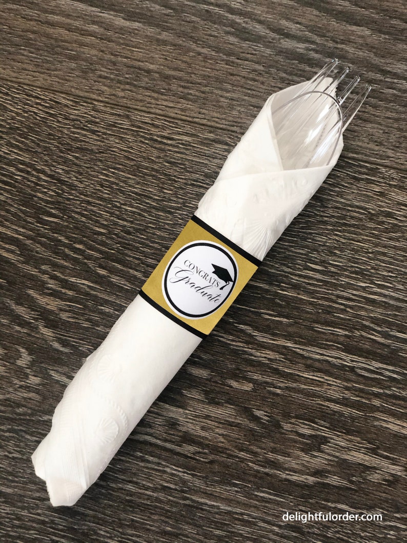 Puede incluir: Una servilleta de papel blanca envuelta alrededor de un tenedor y una cuchara de pl&aacute;stico transparentes con un anillo de servilleta de fiesta de graduaci&oacute;n dorado y negro que dice "Congrats Graduate".