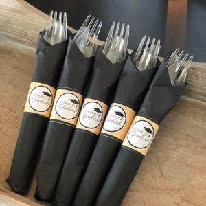 Puede incluir: Servilletas negras con un gorro de graduaci&oacute;n dorado y etiquetas de "Congrats Graduate" envueltas alrededor de tenedores de pl&aacute;stico transparentes.