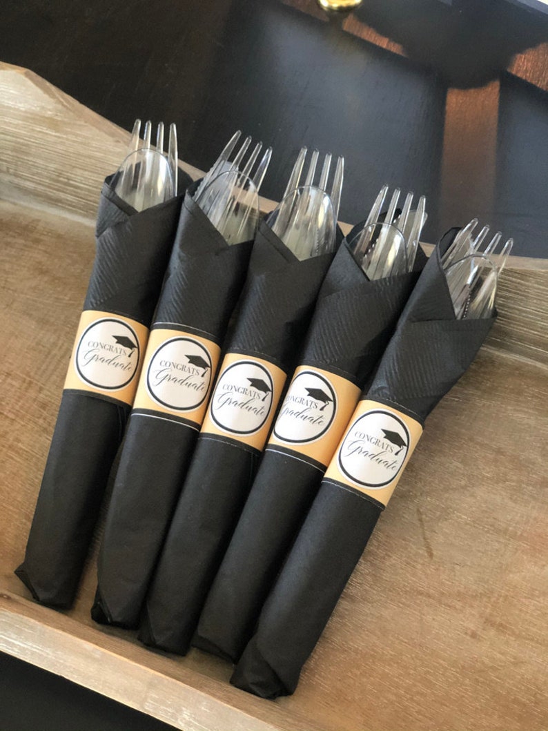 Puede incluir: Servilletas negras con un gorro de graduaci&oacute;n dorado y etiquetas de "Congrats Graduate" envueltas alrededor de tenedores de pl&aacute;stico transparentes.