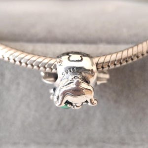 Puede incluir: Un charm de plata con dise&ntilde;o de perro, con una peque&ntilde;a piedra verde. El charm est&aacute; marcado con "S925" y est&aacute; en una cadena de plata. El charm es probablemente para una pulsera o collar.
