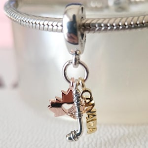 Op de afbeelding: Een zilveren bedel met drie hangers. Een esdoornblad van roségoud, een zilveren hockeystick en gouden letters met de tekst "CANADA". De bedel is bevestigd aan een zilveren armband.