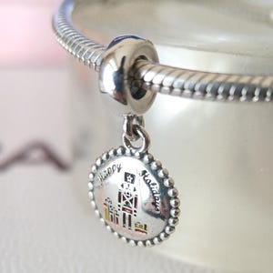 May include: A silver charm with a festive design. The charm features a nutcracker soldier and the words "Happy Holidays" in black script. The charm is attached to a silver bracelet.