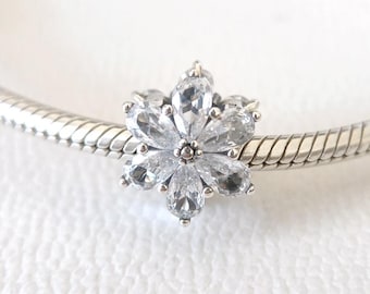 May include: A silver charm with a flower design. The charm features a central cluster of small, round accents surrounded by six clear, teardrop-shaped stones. The charm is attached to a silver bracelet with a textured, snake-like pattern.