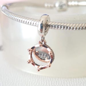 Könnte beinhalten: Ein silberner Charm mit einem roségoldenen Kronen-Design. Der Charm hat die Gravur "QUEEN". Der Charm ist an einem silbernen Armband befestigt. Es ist ein Schmuckstück.