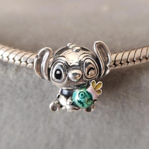 Puede incluir: Un charm de plata con el personaje Stitch de la pel&iacute;cula Lilo & Stitch. El charm muestra a Stitch sosteniendo un objeto verde y turquesa con un lazo rosa. El charm est&aacute; detallado con esmalte negro y est&aacute; en una pulsera de plata.