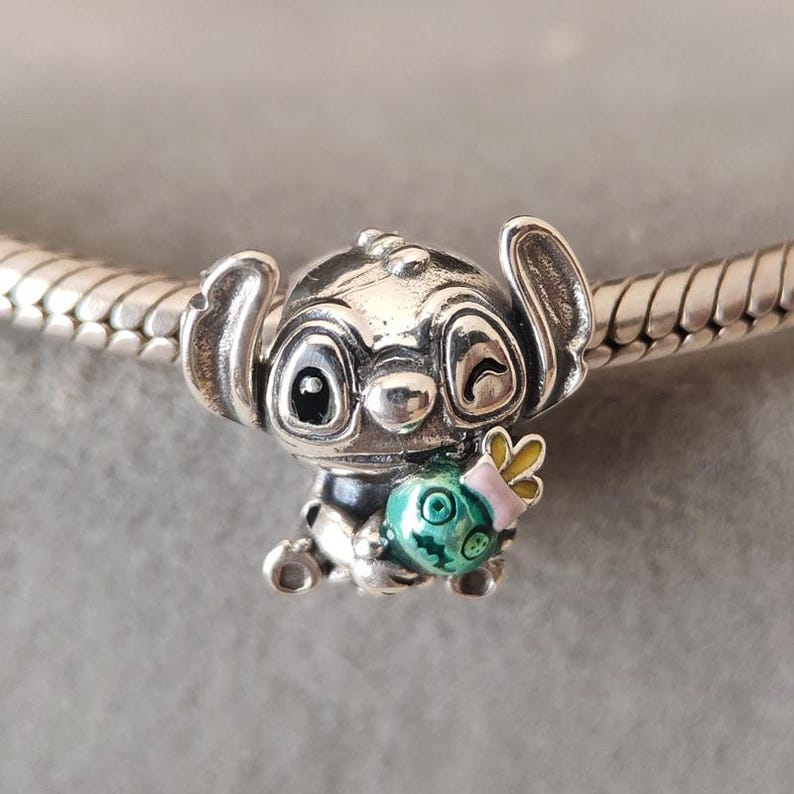 Puede incluir: Un charm de plata con el personaje Stitch de la pel&iacute;cula Lilo & Stitch. El charm muestra a Stitch sosteniendo un objeto verde y turquesa con un lazo rosa. El charm est&aacute; detallado con esmalte negro y est&aacute; en una pulsera de plata.