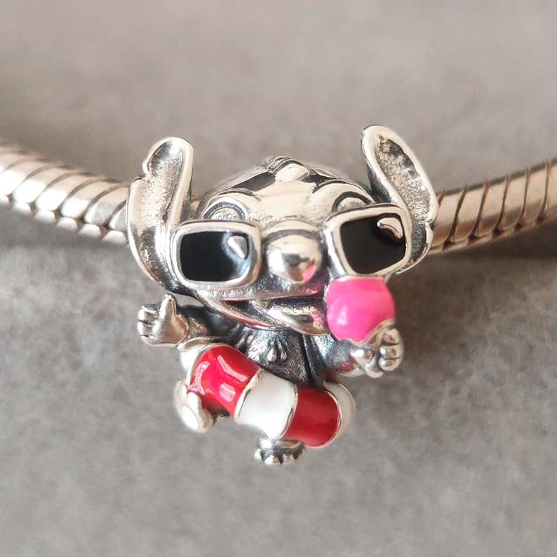 K&ouml;nnte beinhalten: Ein silberner Charm-Anh&auml;nger in Form einer Figur mit gro&szlig;en Ohren, die eine schwarze Sonnenbrille tr&auml;gt, ein leuchtend rosa Eis am Stiel h&auml;lt und einen rot-wei&szlig; gestreiften Rettungsring tr&auml;gt, aufgef&auml;delt auf ein silbernes Schlangenkettenarmband.