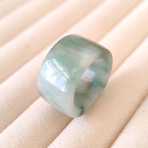 K&ouml;nnte beinhalten: Ein breiter, polierter Jade-Ring mit glattem, abgerundetem Design. Der Ring weist eine Mischung aus hellgr&uuml;nen und wei&szlig;en Farbt&ouml;nen auf, wodurch ein Marmoreffekt entsteht. Der Jade-Ring ist ein Schmuckst&uuml;ck.