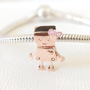 May include: A rose gold charm in the shape of a small person with a pink bow. The charm has a heart on the front and is attached to a silver bracelet. The charm has a small hat and arms and legs.
