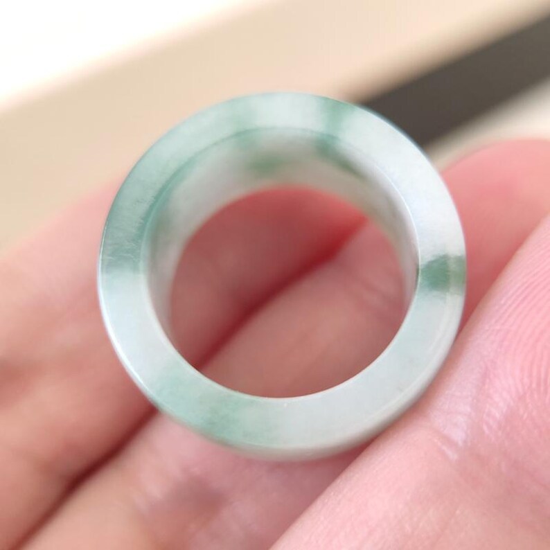 K&ouml;nnte beinhalten: Ein Jade-Ring mit glatter, polierter Oberfl&auml;che. Der Ring hat eine hellgr&uuml;ne und wei&szlig;e Farbgebung mit subtilen Tonvariationen. Der Ring ist ein massives Band, das die nat&uuml;rliche Sch&ouml;nheit des Jades zeigt.