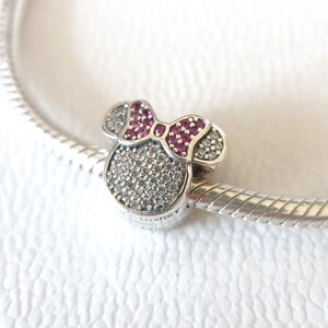 Op de afbeelding: Een zilveren bedel in de vorm van een muizenkop met een strik. De strik is diep roze van kleur en de kop is bedekt met kleine, sprankelende steentjes. De bedel is bevestigd aan een zilveren armband.