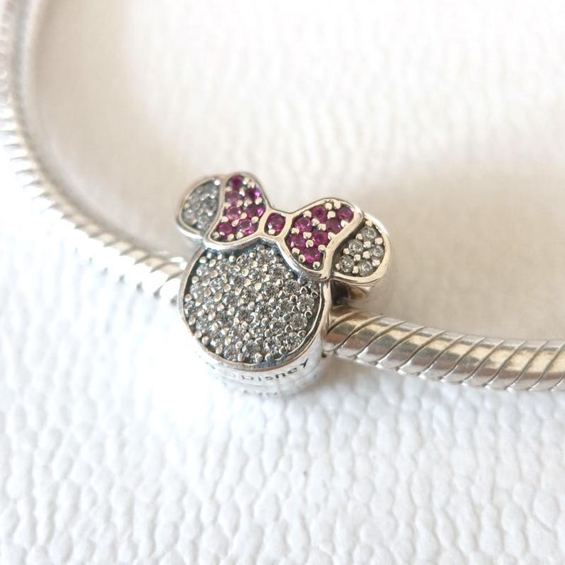 Op de afbeelding: Een zilveren bedel in de vorm van een muizenkop met een strik. De strik is diep roze van kleur en de kop is bedekt met kleine, sprankelende steentjes. De bedel is bevestigd aan een zilveren armband.