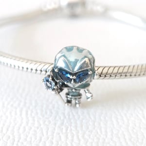 Puede incluir: Un charm de plata con un diseño de robot azul y detalles de piedras preciosas azules. El robot tiene un casco detallado y está unido a una pulsera de plata. Este charm es un accesorio de joyería.