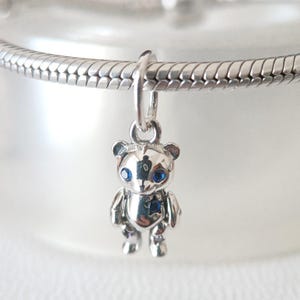 Pode incluir: Um berloque de urso de pelúcia prateado com olhos de pedra azul e uma gema em forma de estrela no peito. O berloque está suspenso de uma corrente prateada, mostrando um design detalhado e polido. As características do urso são claramente definidas.