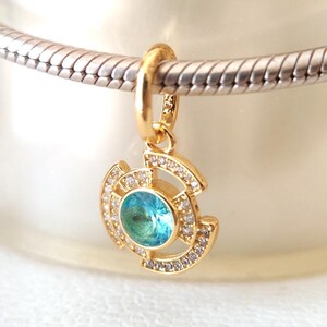 May include: A gold-colored charm with a central, round, light blue gemstone. The charm is surrounded by smaller, clear gemstones and is suspended from a silver-colored bracelet. The charm has a unique, geometric design.