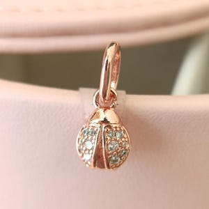 Può includere: Ciondolo a forma di coccinella in oro rosa con anello per bracciale o collana. Il corpo della coccinella è rotondo e ornato da piccole pietre preziose azzurro chiaro. Il ciondolo è esposto su uno sfondo rosa tenue.
