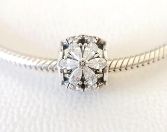 May include: A silver charm with a flower design. The charm features a central flower with clear, sparkling petals and a surrounding decorative pattern. The charm is on a silver bracelet.