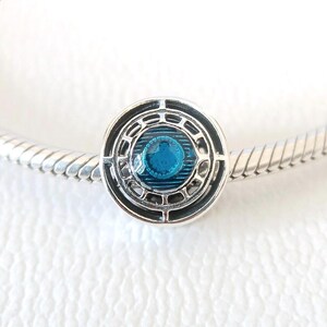 Puede incluir: Un charm de plata con un diseño circular. El charm presenta una piedra azul central rodeada por un patrón calado detallado. El charm está unido a una pulsera de plata, creando una joya.