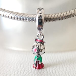 Könnte beinhalten: Ein silberner Charm mit einer kleinen, detaillierten Figur. Die Figur hat einen silbernen Kopf mit einer roten Blume und einem grün-roten Outfit. Der Charm ist an einem silbernen Armband befestigt.