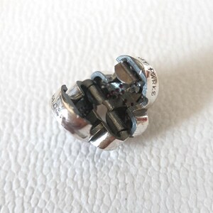 Op de afbeelding: Een zilverkleurige bedel met een bloemmotief. De bedel is open en onthult interne mechanismen en kleine rode accenten. Het woord "PARKS" is gegraveerd op de zijkant. De bedel ligt op een witte ondergrond.