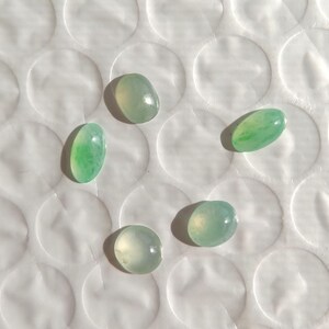May include: Five oval-shaped, polished jade stones in varying shades of green. The stones are arranged in a circular pattern on a white, textured surface. The jade stones have a smooth, glossy finish.