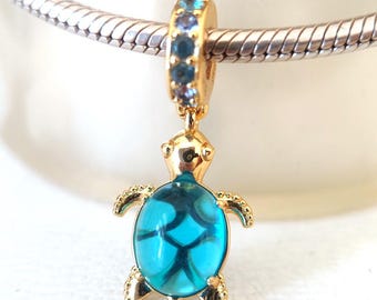 Puede incluir: Un charm de color dorado con un diseño de tortuga turquesa. La tortuga tiene un caparazón azul con dibujos y está suspendida de un anillo dorado adornado con piedras preciosas azules. El charm está diseñado para ser adjuntado a una pulsera o collar.