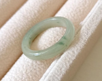 May include: A light green jade ring with a smooth, polished surface. The ring has a circular shape and features subtle variations in color, with darker green inclusions. The ring is set against a soft, off-white fabric background.