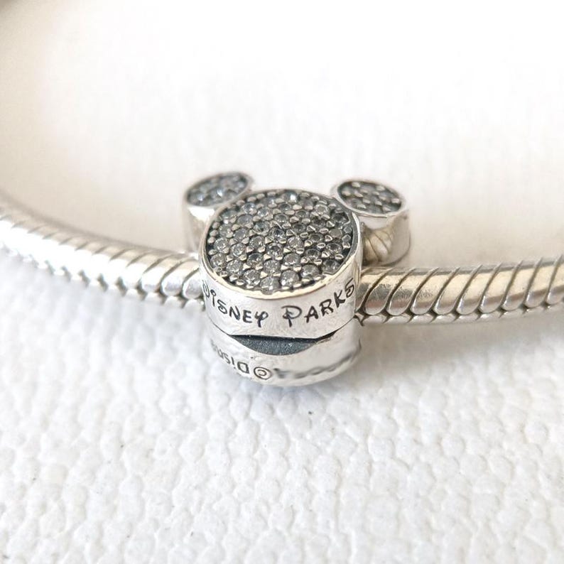 Op de afbeelding: Een zilveren bedel in de vorm van Mickey Mouse's hoofd. Het centrale deel van de bedel is bedekt met kleine, sprankelende steentjes, met twee kleinere, afgeronde steentjes erboven. De woorden "Disney Parks" zijn aan de zijkant gegraveerd.
