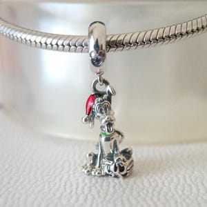May include: A silver charm featuring a cartoon dog wearing a red Santa hat and a green collar. The charm is attached to a silver bracelet. The dog is sitting next to a wrapped gift.