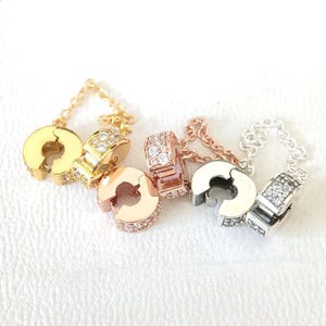 May include: An assortment of charm clasps in gold, rose gold, and silver tones. Each clasp features a circular design with a hinged opening and is adorned with sparkling clear gemstones. Delicate chain details are also visible.